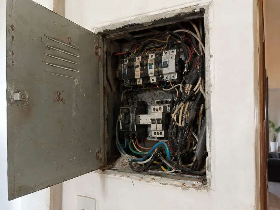 Quadro de luz antigo, com fios emaranhados, fita isolante e disjuntores sem identificação
